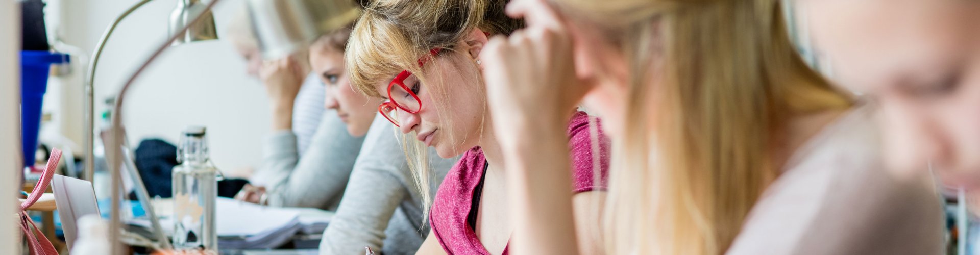 Mehrere Studierende, die in einer Reihe an einem langen Tisch sitzen und über Bücher gebeugt sind oder am Laptop arbeiten. Einige sind unscharf zu erkennen, den Bildmittelpunkt bildet eine blonde Frau mit auffällig roter Brille.