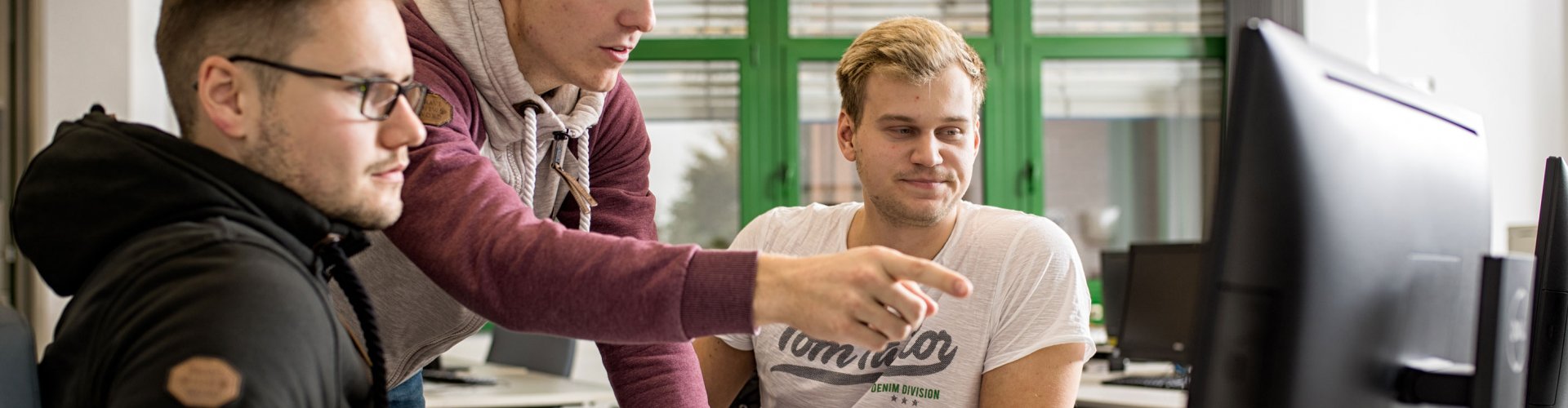 Drei Studenten schauen gemeinsam auf einen Computerbildschirm. Der in der Mitte stehende zeigt den anderen beiden, die sitzen, etwas.