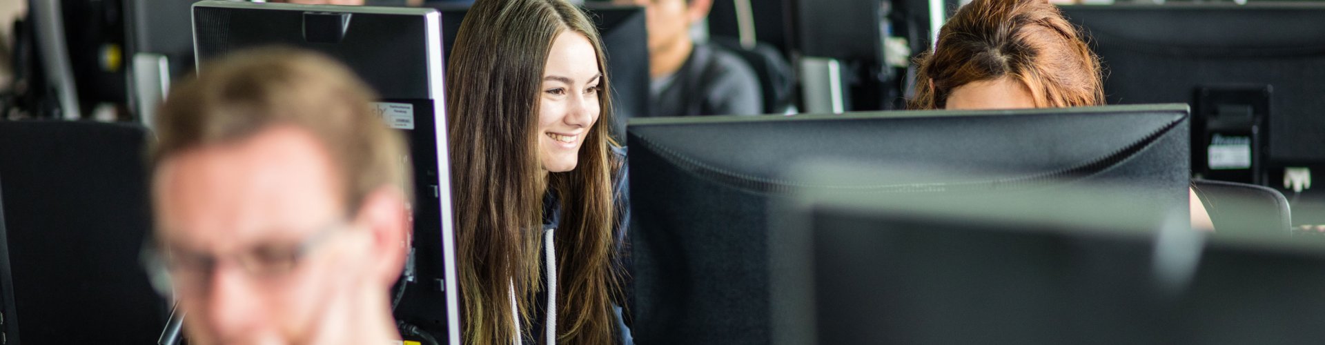 Blick auf ein Computerlabor, in dem mehrere Studierende an Rechnern arbeiten. In der ersten Reihe ist unscharf ein blonder Student zu erkennen, im Fokus in der Reihe hinter ihm eine dunkelhaarige, lachende Studentin.