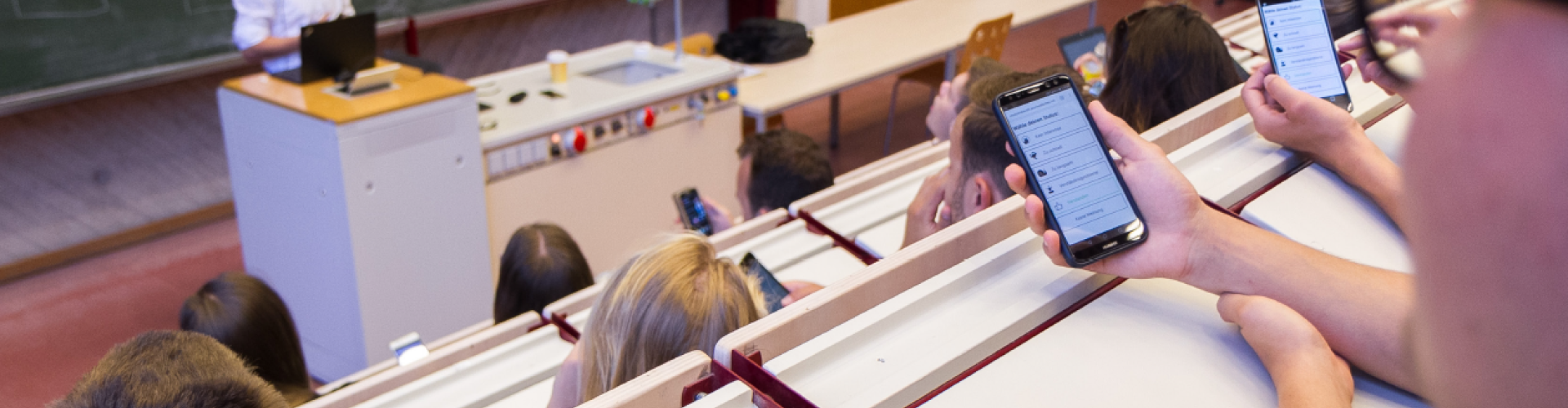 Hörsaal mit Dozent im Hintegrund. Im Vordergrund Studierende, die ihre Smartphones in den Händen halten.