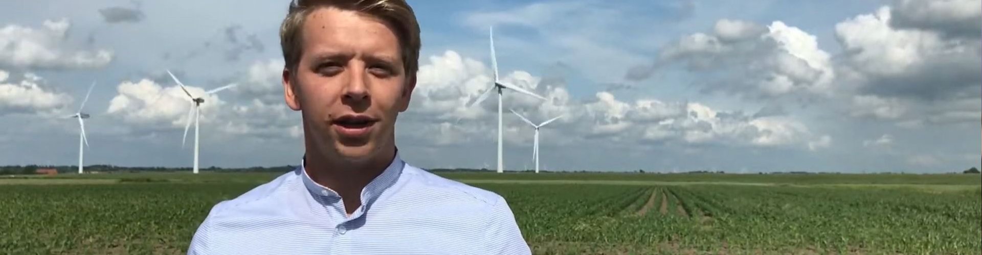 Junger Mann mit kurzen blonden Haaren und hellbaluem Hemd steht auf einer Wiese. Im Hintergrund Wolken am Himmel und Windräder.