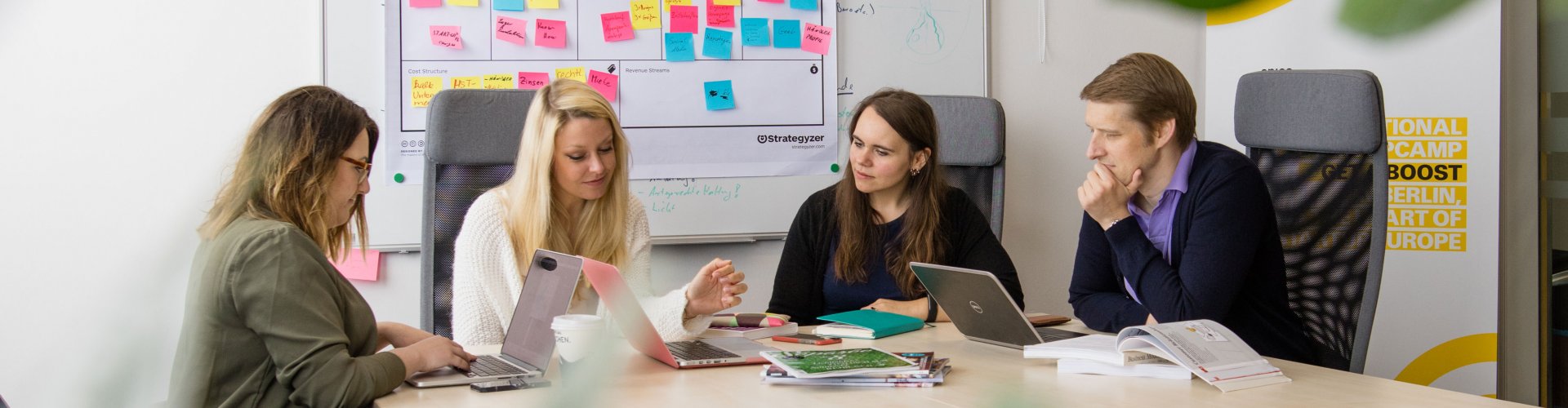 Eine Gruppe von Menschen sitzt um einen Arbeitstisch, hinter ihnen an der Wand hängt ein Whiteboard voller bunter Post Its, vor ihnen auf dem Tisch Laptops, Bücher und Stifte.