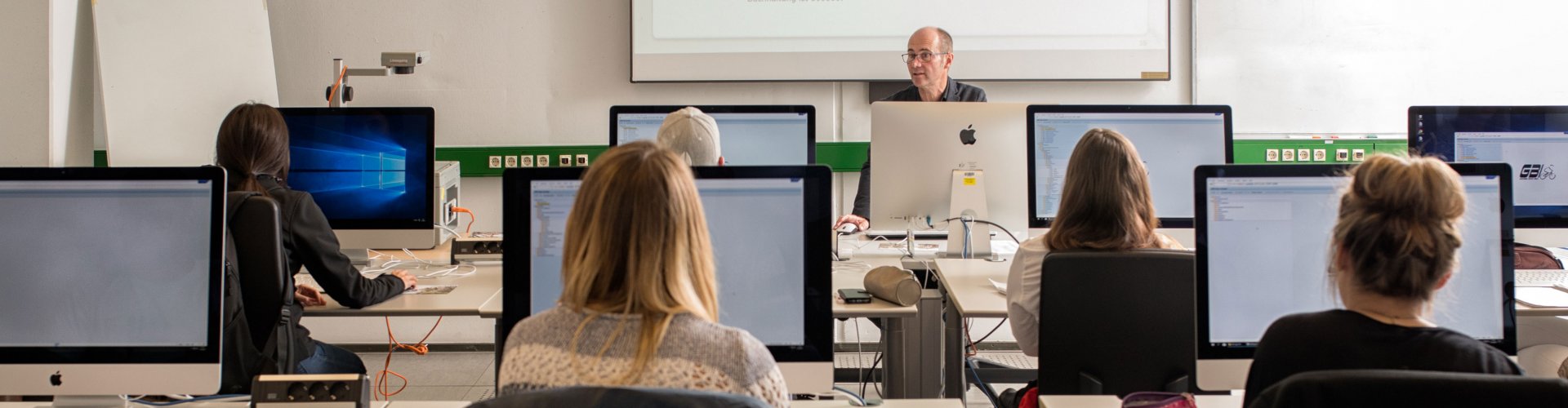 Blick von einen Studierenden auf seine Mitstudierenden, die vor Ihren PC sitzen und einem Dozenten zuhören