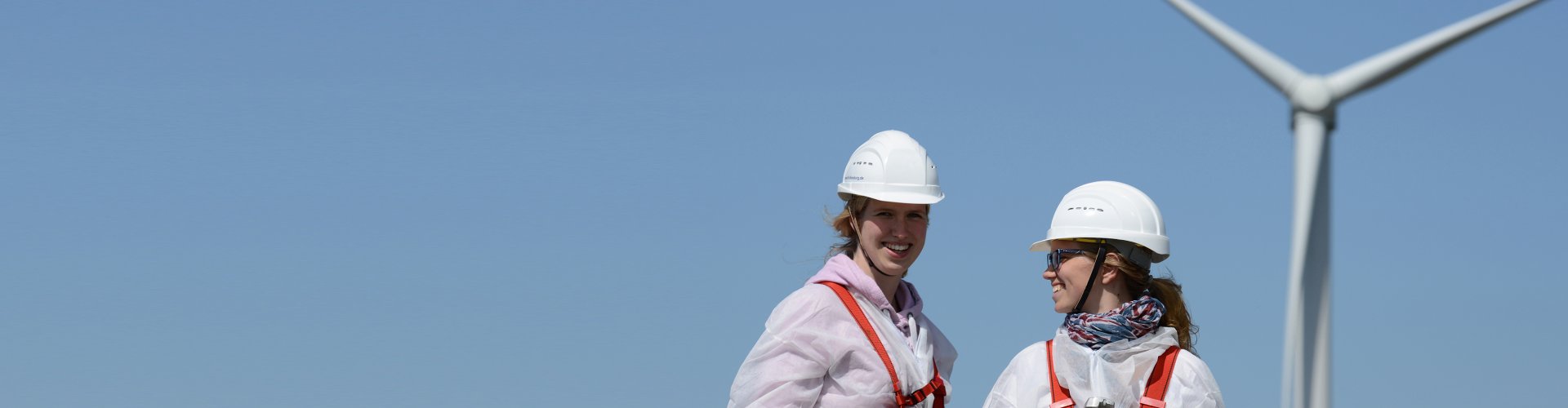 Zwei Frauen mit weißen Helmen stehen vor einer Windkraftanlage