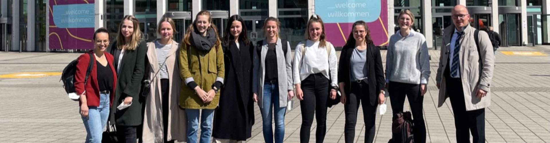 Eine Gruppe Studierender und ein Lehrender posieren vor der Messe Berlin. Am Gebäude hängt ein großes Plakat zur Messe.