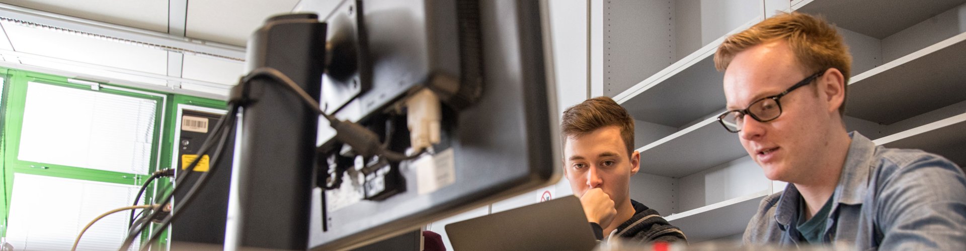 Zwei junge Männer an einem Schreibtisch. Sie blicken auf den Computer vor ihnen.