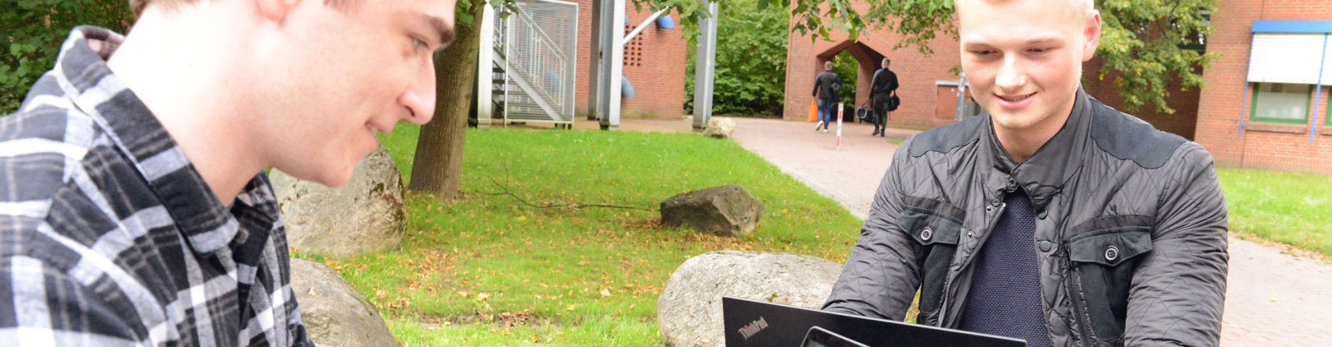 Zwei junge Männer sitzen draußen an einem Tisch und schauen auf Laptops, die vor ihnen stehen.