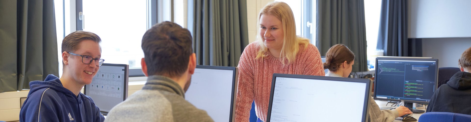 Studierende in einem Seminarraum. Zwei Studenten schauen auf Bildschirme, eine Studentin steht dahinter. Sie unterhalten sich.