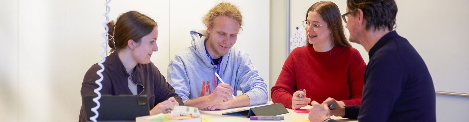 Studierende und ein Dozent stehen um einen Tsich. Sie halten Stifte, einer macht Notizen auf dem Tablet.