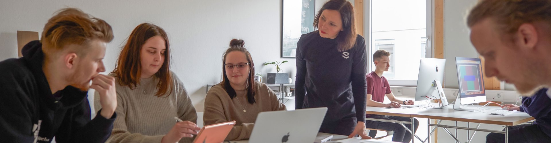 Studierende arbeiten in einem hellen, freundlichen Raum an Laptops. Eine DSozentin steht daneben und hört interessiert zu.