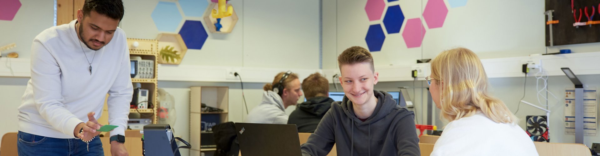 Studierende in einem freundlichen Arbeitsraum, sie sitzen an Tischen, halten Platinen in der Hand und reden.