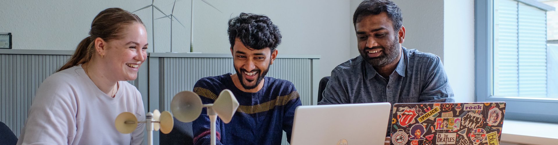 Drei Studierende arbeiten an Laptops, hinter und vor ihnen Modelle von Windkraftanlagen. Sie lachen.