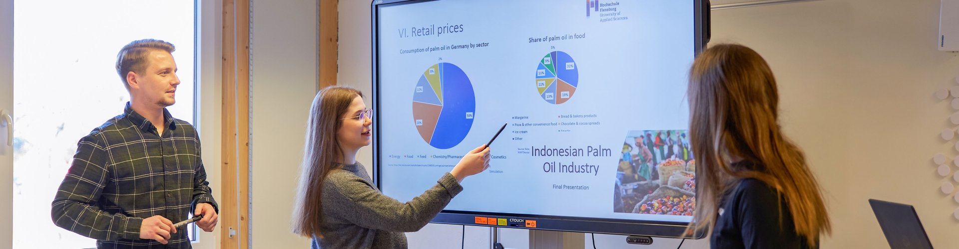 Drei Studierende stehen um einen Bildschirm, auf dem Diagramme zu erkennen sind. Eine Studentin zeigt etwas.