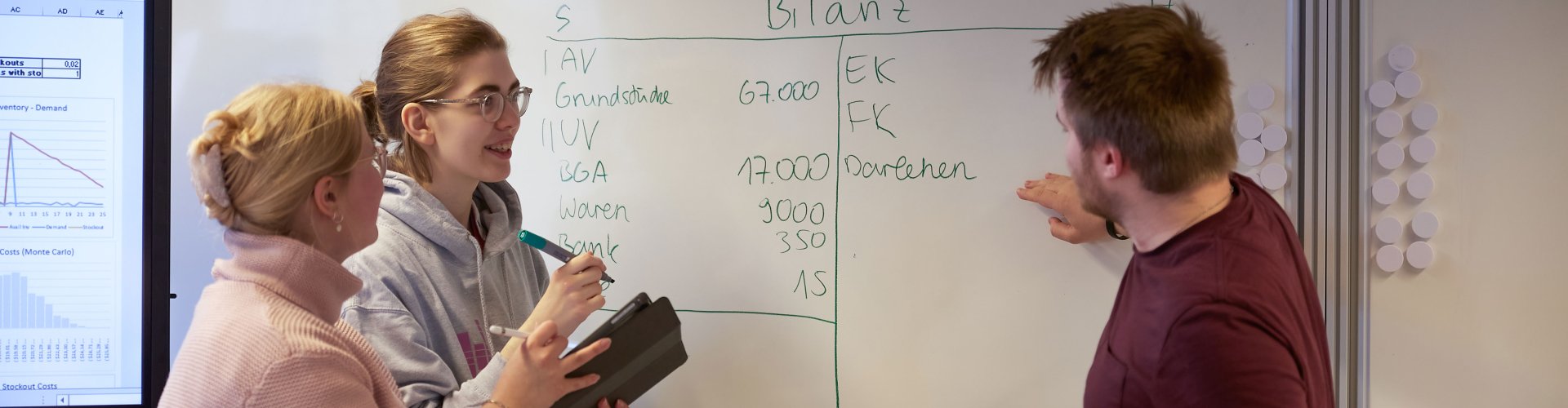 Drei Studierende an inem Whiteboard, es ist u.a. das Wort "Bilanz" zu erkennen.