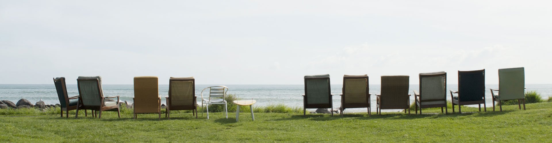 Auf einer Wiese vor dem Meer sind mehrere Sessel in einer Reihe aufgestellt. Wenn man sich hineinsetzen würde, hätte man einen Blick auf das Meer.