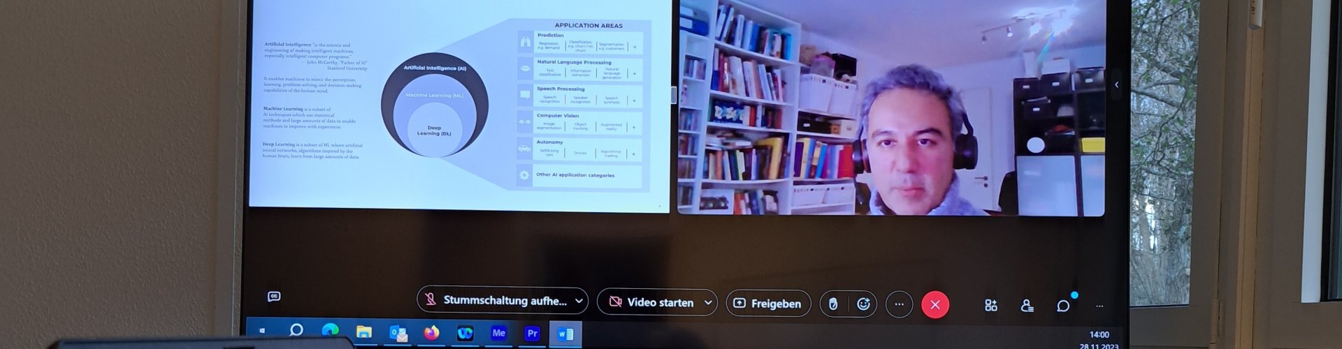 Auf einem großen Monitor ist ein Splitscreen mit einer Präsentation und einem Mann mittleren Alters zu sehen, der in die Kamera spricht. Die Präsentation hat den Titel "AI Use Cases in Media". Im Vordergrund ein Tisch mit einem aufgeklappten Laptop, einem Schreibblock mit Notizen und einer Kaffetasse mit dem Logo der Hochschule Flensburg.