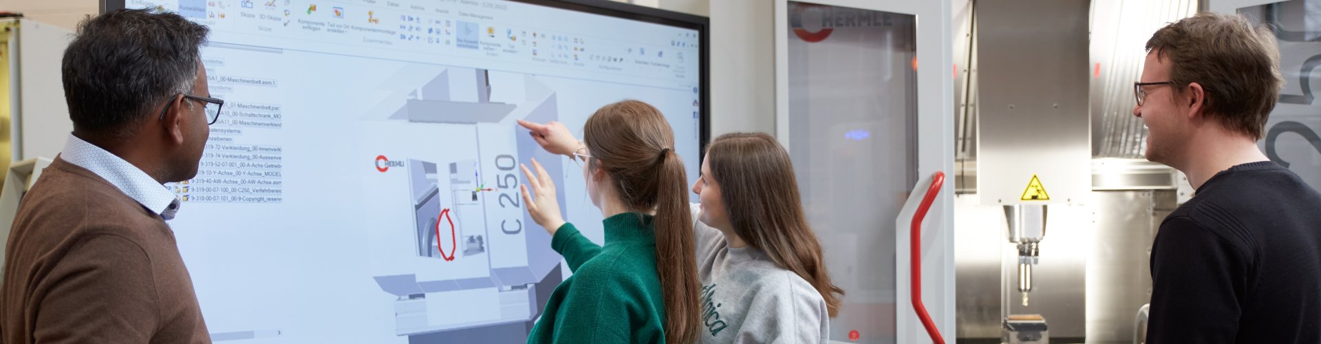 Zwei Studentinnen stehen an einem großen Monitor und zeigen auf die dort dargestellte Grafik mit verschiedenfarbigen eckigen Flächen und der Angabe C250. Ein Dozent steht links, ein Student links neben ihnen und blicken ebenfalls auf den Bildschirm.