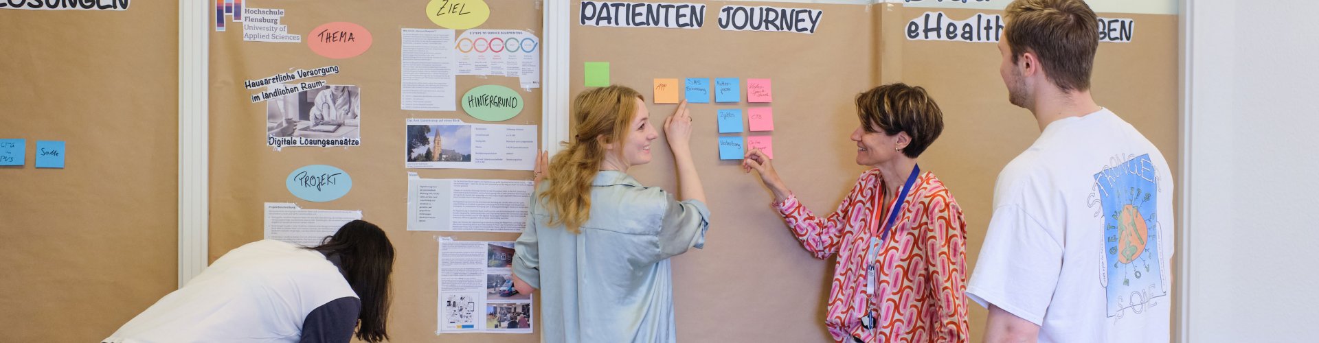 Studierende und Dozentin vor einer Pinnwand. Sie sind im Gespräch. Zu erkennen sind Wörter wie "eHealth" und "Patienten Journey".