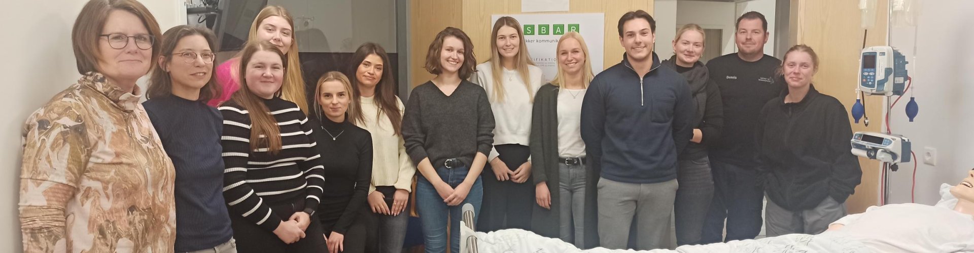 Eine Gruppe von jungen Menschen steht rund um ein Krankenhausbett, in dem eine Modellpuppe liegt. An der Wand hängt ein Monitor, neben dem Bett ein Tropf und an der Wand hinter den Studierenden hängt ein Poster mit Aufschrift in dänischer Sprache.