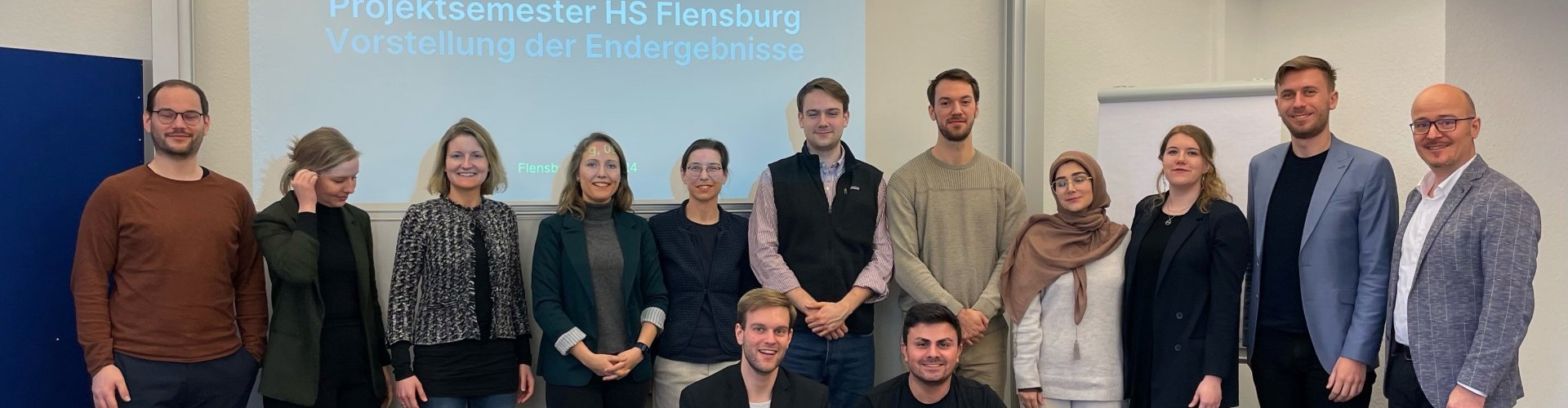 Eine Gruppe von überwiegend jungen Menschen steht in einer Reihe und lächelt in die Kamera, zwei junge Männer knien davor. Im Hintergrund ist die Projektion einer PPT-Folie zu sehen, auf der auf blauem Hintergrund das Logo der Asklepios-Gruppe zu sehen ist, darunter der Schriftzug: "Projektsemester HS Flensburg. Vorstellung der Endergebnisse".