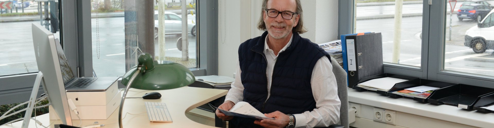 Ein älterer Mann mit schulterlangen grauen Haaren sitzt an einem Schreibtisch und lacht in die Kamera. Durch die Fenster im Hintergrund sieht man eine Straße und den Flensburger Hafen.