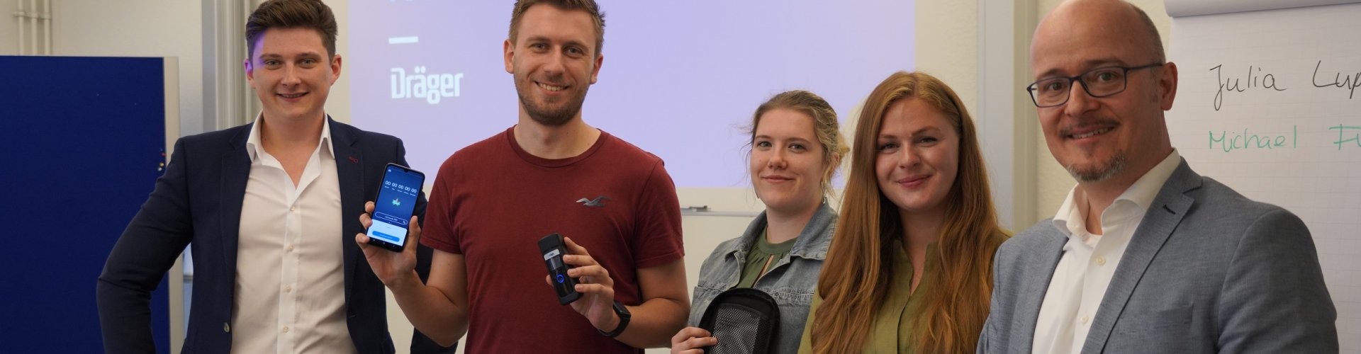Drei Studierende und zwei Männer lächeln in die Kamera, im Hintergrund eine Projektion mit der Überschrift "Technology for Life - Dräger". Einer der Studierendne hält ein Smartphone und ein Messgerät, eine andere ein Schutzcase.