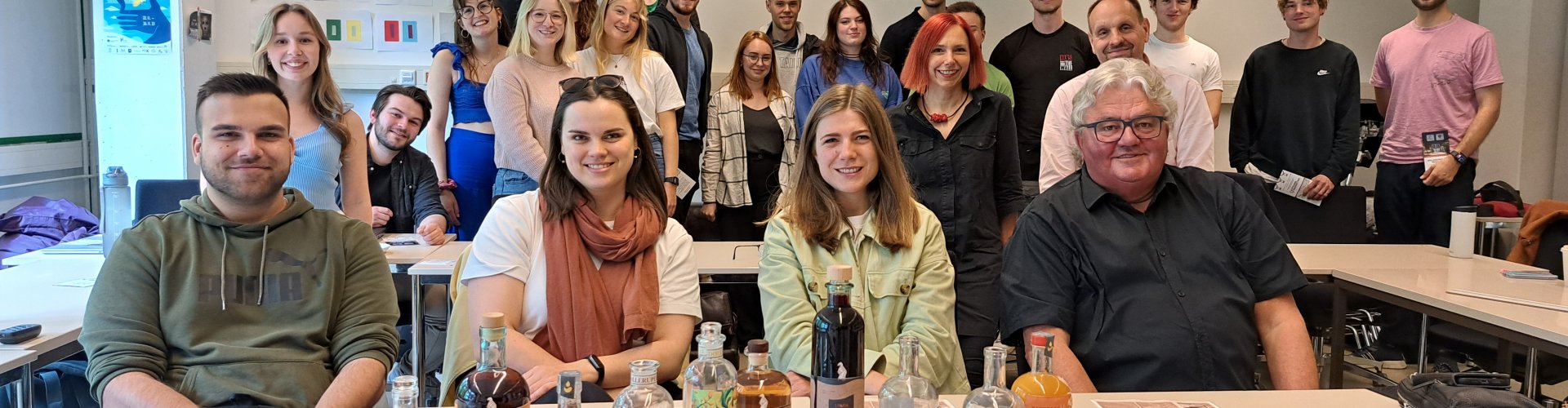 Zwei Männer und zwei Frauen sitzen in einem Seminarraum, vor ihnen ist eine Reihe von Glaschflaschen mit bunten Etiketten aufgebaut. Hinter ihnen steht eine große Gruppe von Studierenden.