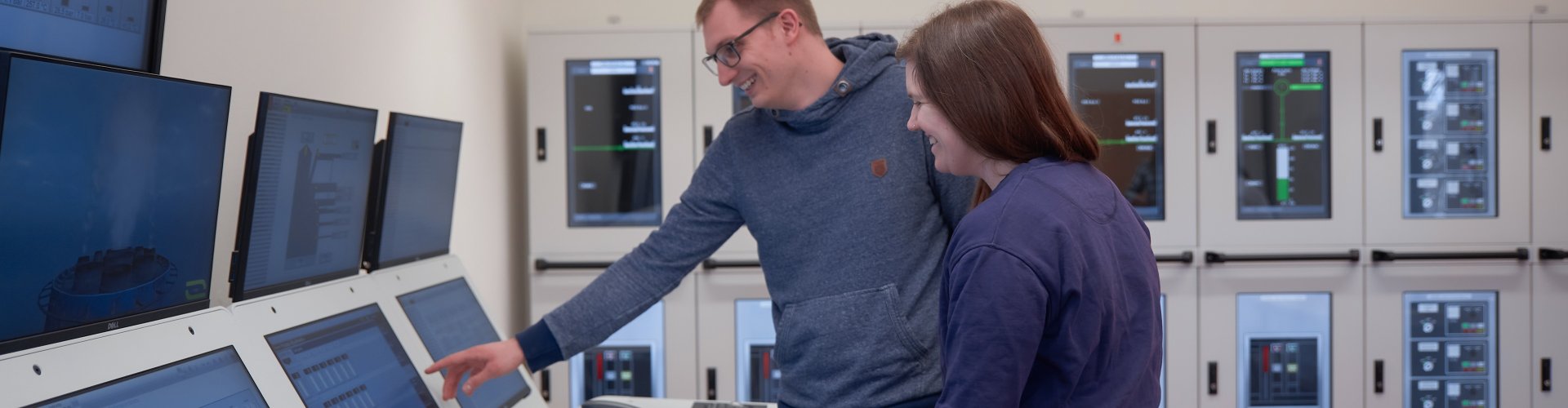 Ein Student und eine Studentin stehen in einem Raum mit vielen Monitoren, auf denen schiffsbetriebstechnische Anzeigen zu sehen sind.