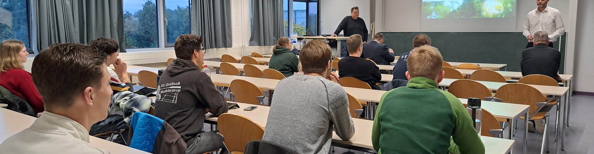 In einem Seminarraum sitzen Studierende in Tischreihen. Vorne stehen zwei Männer, hinter ihnen wird eine Präsentation an die Wand projeziert mit dem Text: Green Process Engineering. GPE-Firmenpräsentation & Projekt: Erneuerung Lufterhitzer