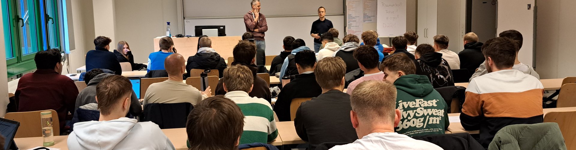 In einem Seminarraum sitzen Studierende und Schüler*innen in Tischreihen, vorne stehen ein Dozent und ein Lehrer und sprechen zur Gruppe.