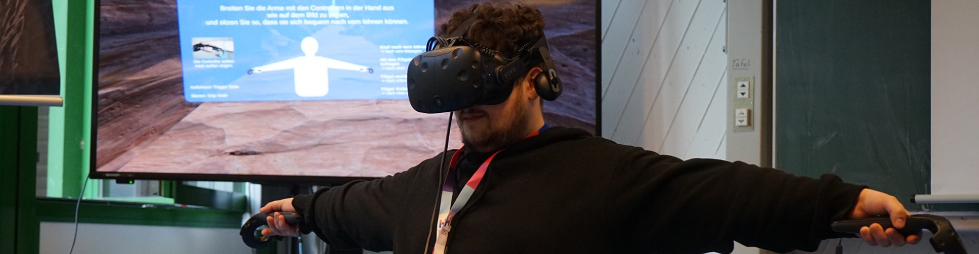 Ein junger Mann trägt eine VR-Brille, hat zwei Controller in den Händen und streckt die Arme zu beiden Seiten aus. Im Hintergrund sieht man einen Bildschirm, auf dem eine virtuelle Umgebung abgebildet ist.