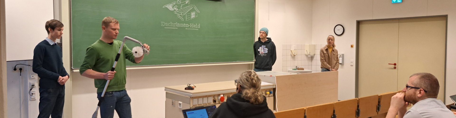 Eine Gruppe von Studierenden steht vorne in einem Vorlesungssaal und hält eine Präsentation. Einer der Studierenden hält ein elekrtisches Gerät mit Teleskoparm in der Hand, an der Tafel hinter ihnen ist ein Logo angezeichnet mit Schriftzug "Dachrinnen-Held"