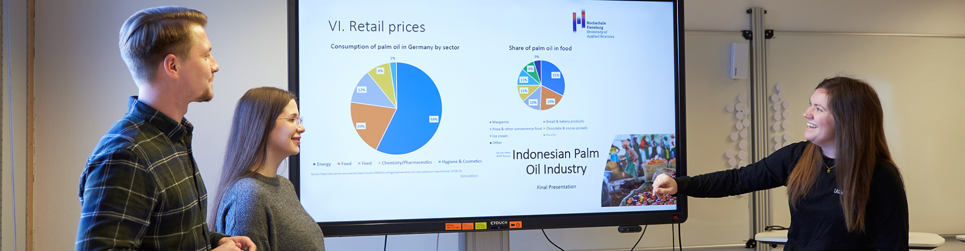 In einem Seminarraum stehen ein Student und zwei Studentinnen vor einem großen Monitor, auf dem verschiedene Tortendiagramme zu sehen sind - neben einem steht in größerer Schrift "Indonesian Palm Oil Industry".
