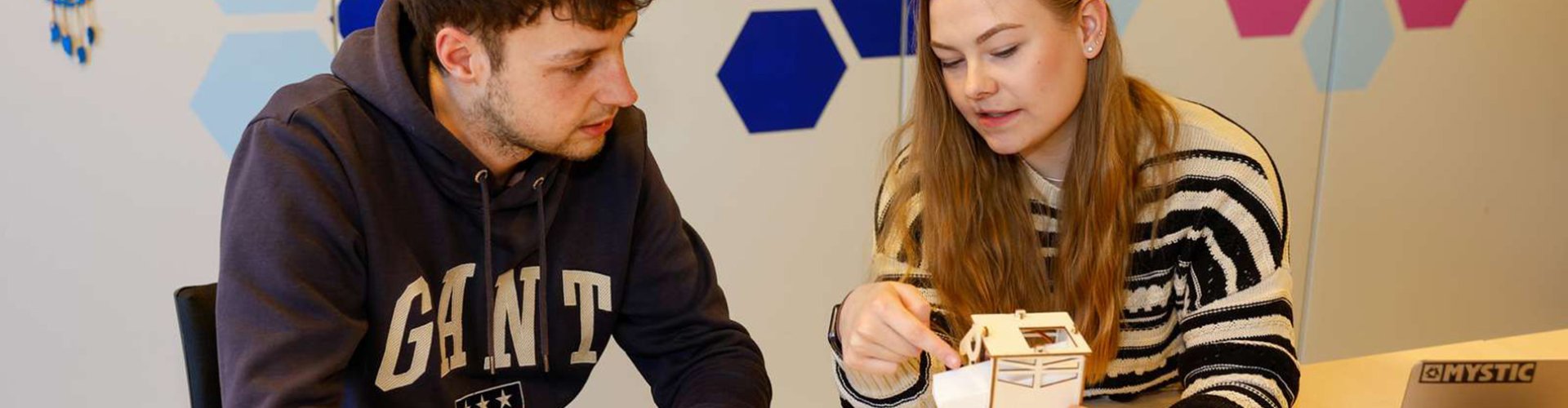Ein junger Mann und eine junge Frau sitzen an einem Tisch, die Frau hält einen kleinen, selbstgebauten Roboter in der Hand und erklärt dem Mann etwas dazu.