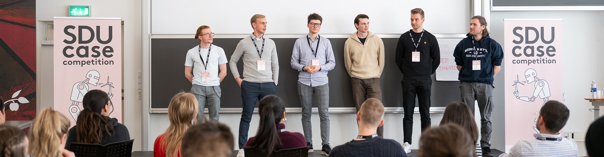 Eine Gruppe von Studierenden steht in einem großen Raum vor einem Publikum und hält einen Vortrag, neben ihnen stehen Roll-Ups von der SDU Case Competition.
