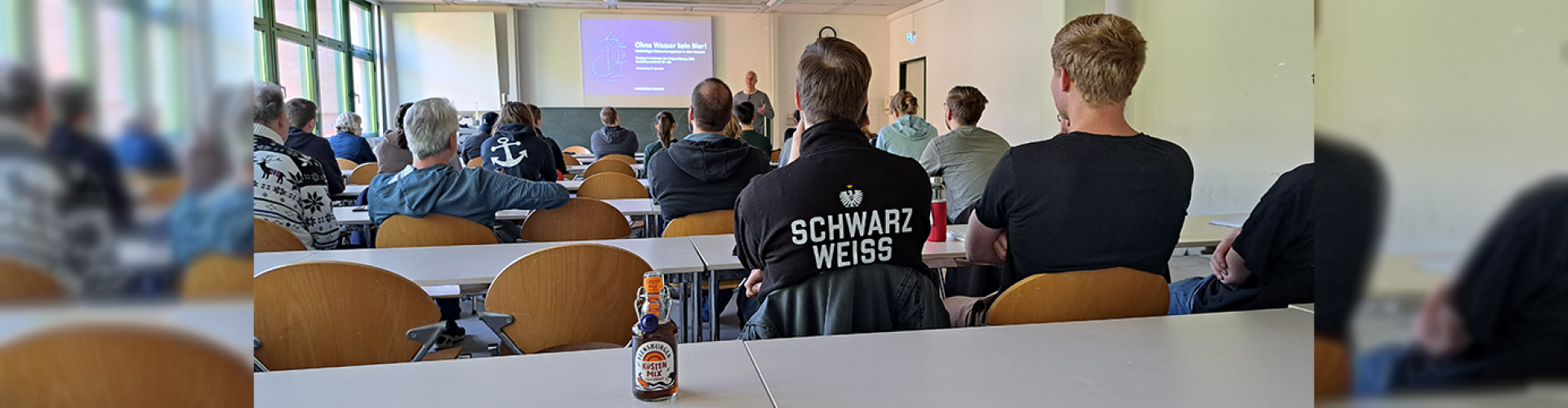 In einem Seminarraum sitzen viele Studierende, vorne steht ein Mann und hält einen Vortrag, seine Präsentation trägt den Titel "Ohne Wasser kein Bier!"