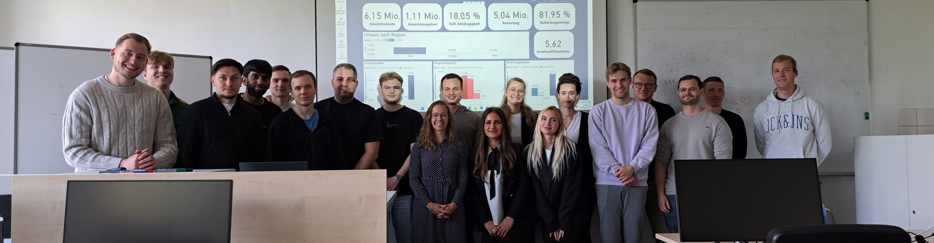 Eine Gruppe von ca. 30 Menschen steht in einem Seminarraum und lächelt in die Kamera, hinter ihnen an der Wand die Projektion einer Präsentationsfolie mit Diagrammen und anderen Daten.