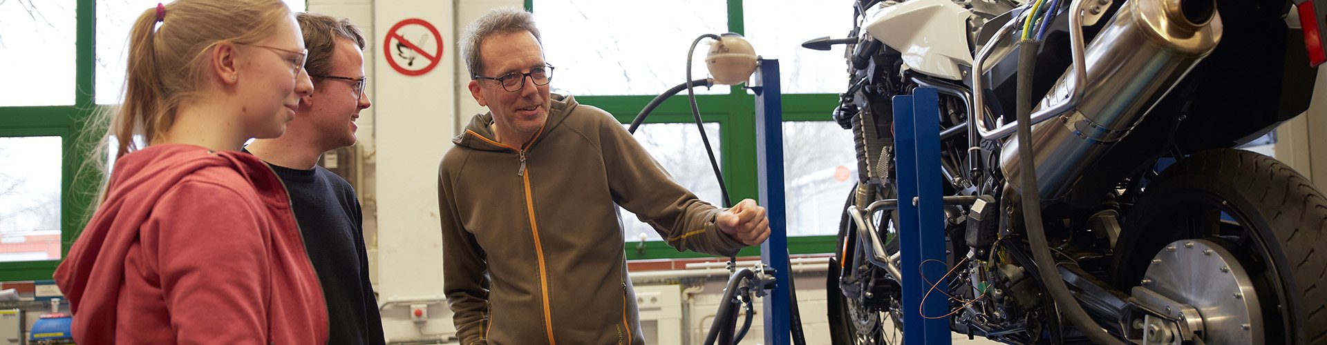 Zwei Studierende und ein Dozent stehen vor einem Motorrad.