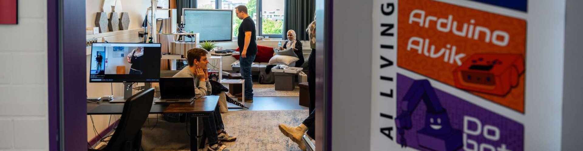 Blick durch eine offene Tür in ein Labor, das mit einem PC-Arbeitsplatz, einem Monitor, einem Regal und einem Sofa ausgestattet ist und in dem mehrere junge Menschen sitzen. 