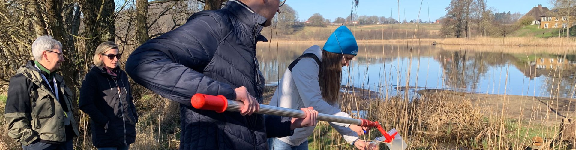 Zwei Menschen entnehmen eine Wasserprobe, im Hintergrund ist der See zu erkennen.