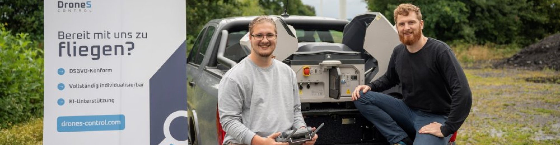 Zwei junge Männer hocken auf der Ladefläche eines Pichup-Trucks, der auf einem Feldweg steht. Einer steuert eine Drohne, neben ihm steht ein Roll-Up mit Text: DroneS Control. Bereit mit uns zu fliegen? DSGVO-konform, vollständig indiovidualisierbar, KI-Unterstützung. drones-control.com