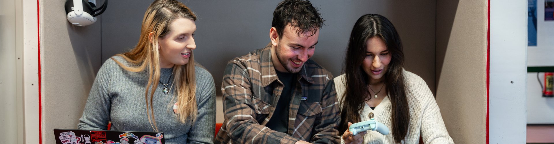 Drei Studierende sitzen in einer Sitzecke und arbeiten gemeinsam an Laptops, zwei haben Spielekonsolen-Controller in der Hand.