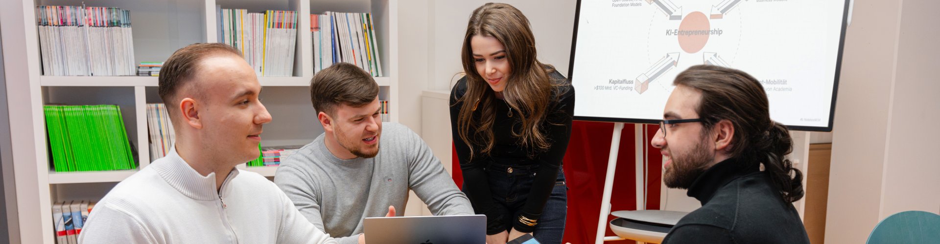 Vier Studierende sitzen um einen Tisch und diskutieren, hinter ihnen ein großer Monitor mit einem Schaubild zu KI-Entrepreneurship.