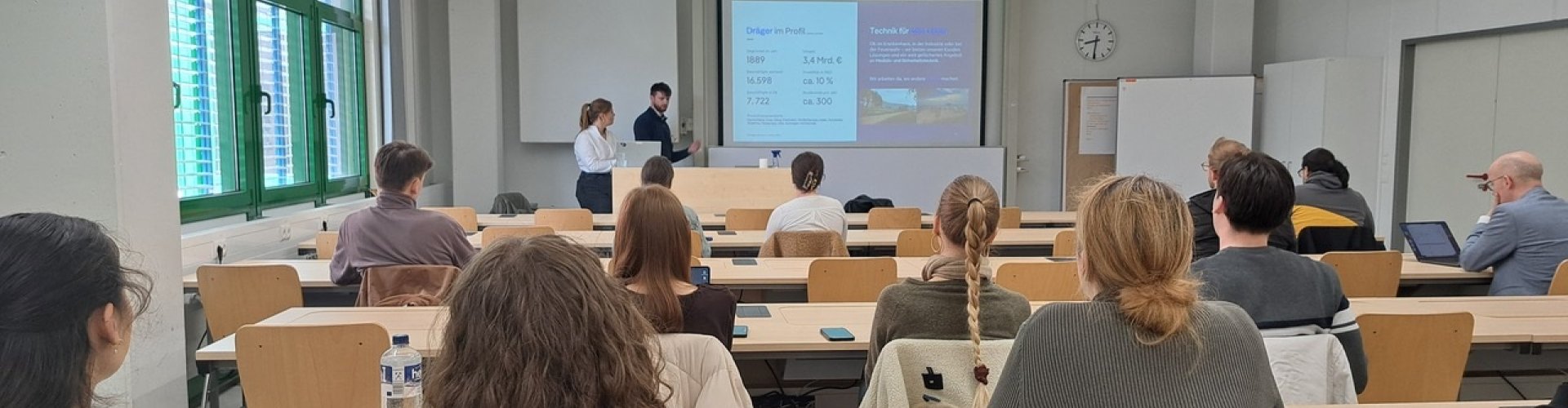 In einem Seminarraum sitzen Studierende und lauschen einem Vortrag, den eine junge Frau und ein junger Mann vorne halten.