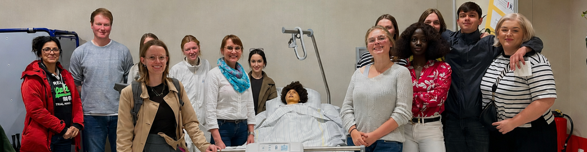 Eine Gruppe von etwa 12 Studierenden steht um ein Pflegebett mit einer Puppe drumherum und lächelt fröhlich in die Kamera.