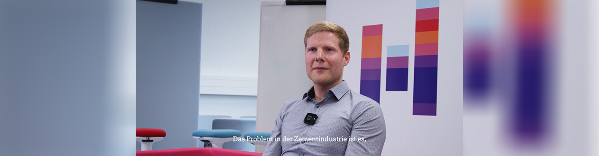 Ein junger Mann im hellblauen Hemd sitzt vor einem Roll-Up mit dem Logo der Hochschule Flensburg und spricht in die Kamera.