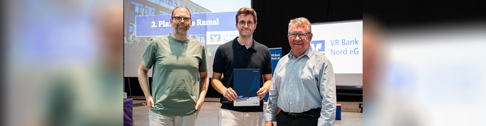 Mats Ramsl, Kai Petersen und Hauke Präger lächeln in die Kamera, hinter ihnen mehrere Leinwände mit dem Logo der VR Bank Nord.