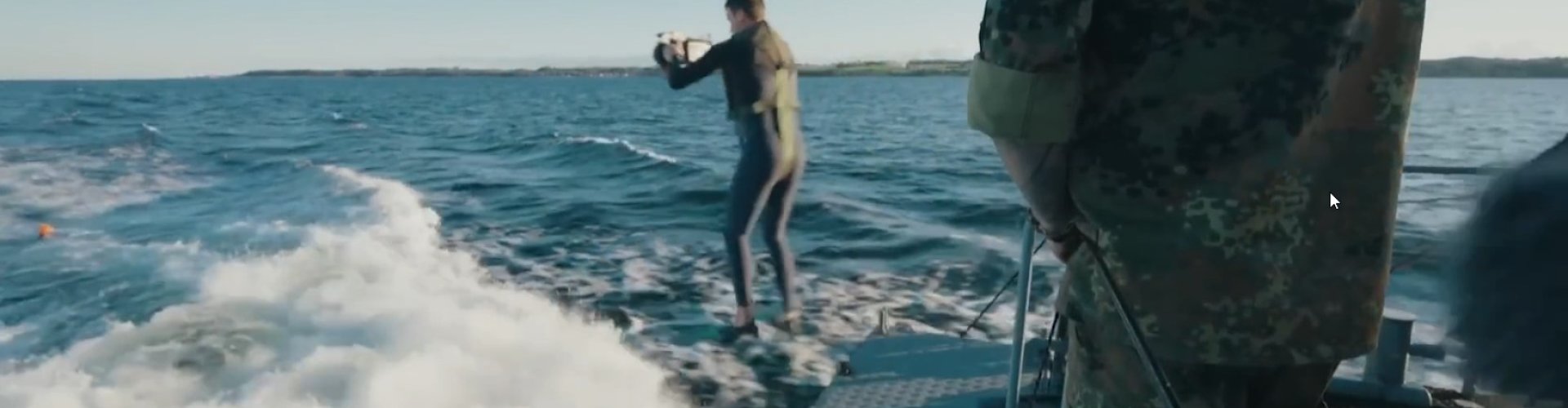 Ein junger Mann im Neoprenanzug mit Filmkamera springt vom Heck eines fahrenden Marine-Schiffes.