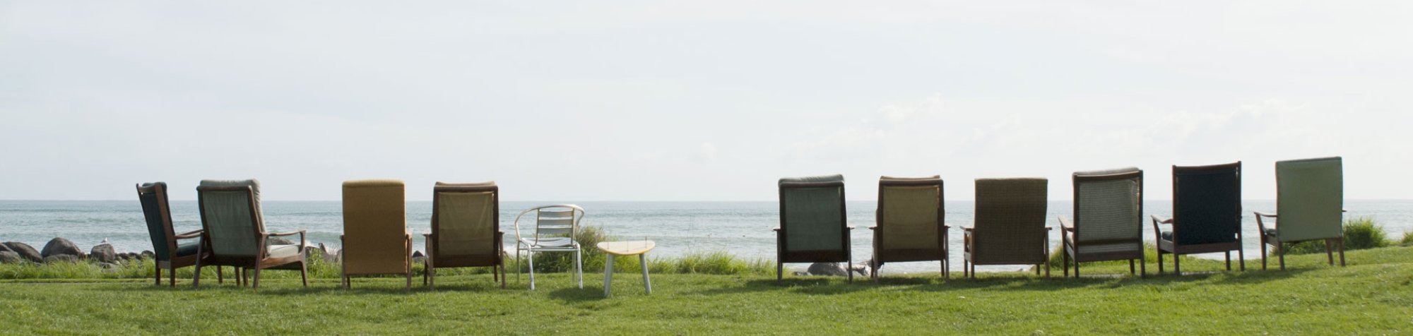 Auf einer Wiese vor dem Meer sind mehrere Sessel in einer Reihe aufgestellt. Wenn man sich hineinsetzen würde, hätte man einen Blick auf das Meer.