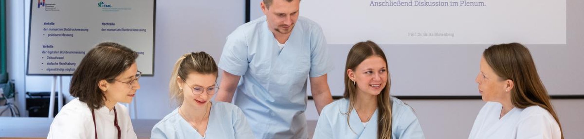 fünf Menschen sitzen am Tisch, einer steht, sie unterhalten sich, haben weiße Kittel an